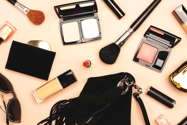 Accessori per cose nere da donna e cosmetici decorativi su sfondo rosa nudo Occhiali da sole rossetto pennello per trucco in polvere Concetto di contenuto della borsa cosmetica per ragazza