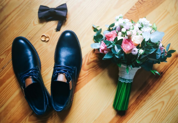 Accessori per il giorno del matrimonio Scarpe da sposo con bouquet sullo sfondo di legno Anelli d'oro e papillon sono nelle vicinanze Cose alla moda