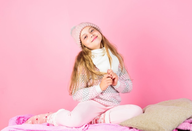 Accessorio di moda invernale. Concetto di accessorio invernale. Ragazza capelli lunghi sognante umore sfondo rosa. Kid sorridente indossare accessorio a maglia. La ragazza del bambino indossa un cappello alla moda lavorato a maglia e un accessorio per sciarpa.