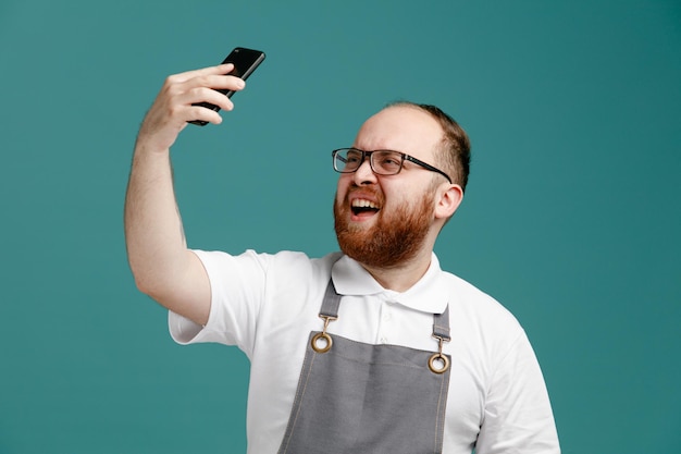 Accigliato giovane barbiere che indossa uniforme e occhiali alzando il telefono cellulare prendendo selfie isolato su sfondo blu