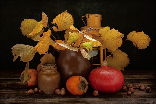 Accogliente natura morta autunnale con foglie gialle, lanterna, zucca, cachi e nocciola su tavola di legno