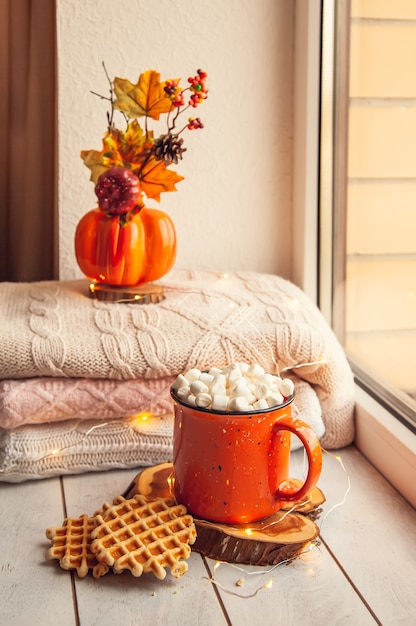 Accogliente natura morta autunnale sul davanzale: caldi maglioni di lana, zucche, foglie d'acero e una tazza di cacao con marshmallow e cialde.