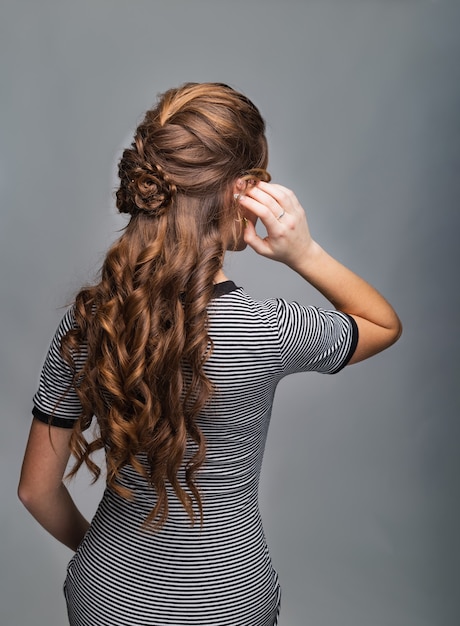 Acconciatura ricci ondulati. Acconciatura su capelli castani rossi donna con capelli lunghi su sfondo grigio. Servizi professionali di parrucchieri. Acconciatura, realizzazione di trecce con forcina.