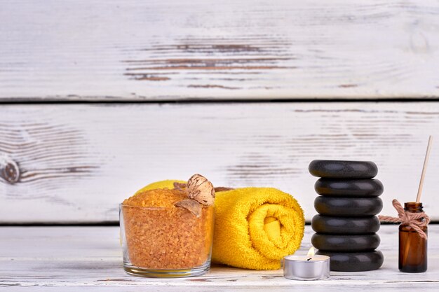 Acessori di trattamento spa still life su fondo di legno bianco. Asciugamano arrotolato giallo con sale himalayano e pila di pietre nere.