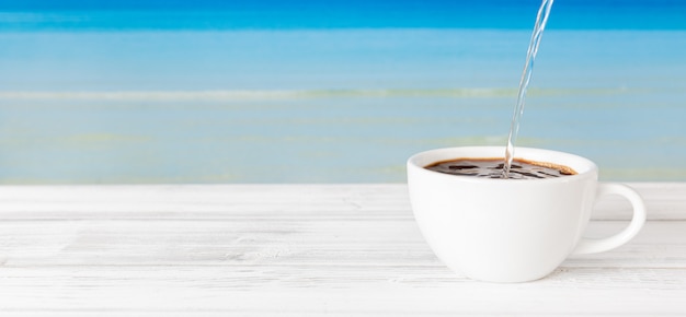Acqua calda di versamento nella tazza di caffè sulla tavola di legno bianca con il fondo blu luminoso del mare