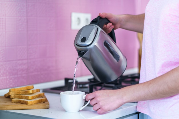 Acqua di ebollizione di versamento della donna da un bollitore elettrico in una tazza per fare tè caldo a casa alla cucina