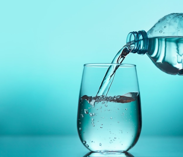 Acqua fresca che versa dalla bottiglia di plastica in un bicchiere su sfondo blu. Bere acqua ogni giorno è importante per una salute ottimale