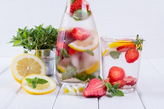 Acqua minerale infusa con fragole, ghiaccio, erbe e foglie di menta su superficie bianca, ricetta detox fatta in casa con acqua di soda.