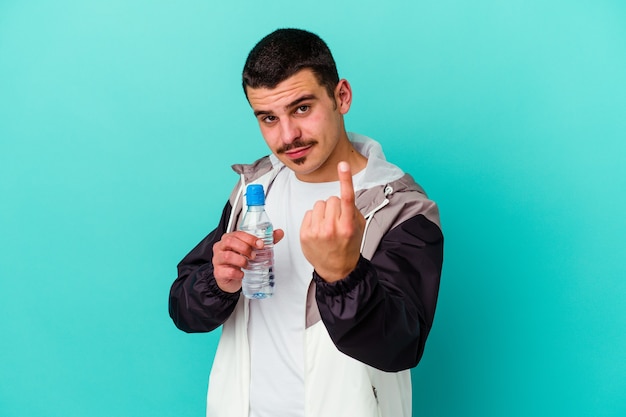 Acqua potabile del giovane uomo caucasico sportivo isolata sull'azzurro che indica con il dito contro di voi come se invitando si avvicinano.