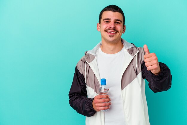 Acqua potabile del giovane uomo caucasico sportivo isolata sulla parete blu che sorride e che alza il pollice in su