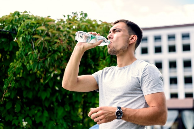 Acqua potabile del giovane uomo europeo dalla bottiglia di plastica su fondo dei cespugli verdi degli alberi il giorno soleggiato luminoso