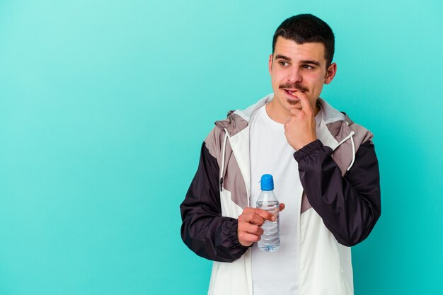 Acqua potabile dell'uomo caucasico sportivo giovane isolata sul pensiero rilassato blu a qualcosa che guarda uno spazio della copia.