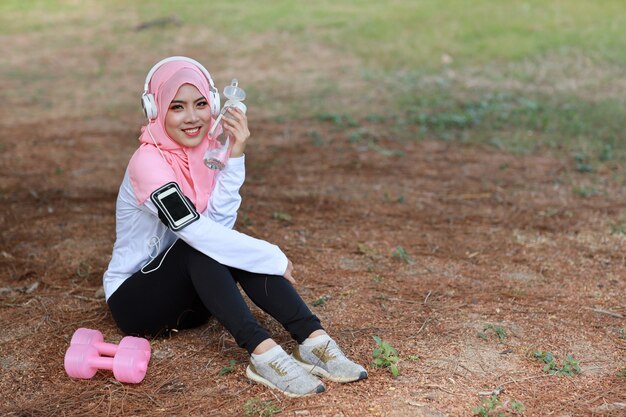 Acqua potabile della bella donna musulmana asiatica dell'atleta di forma fisica dopo l'esercizio. Giovane ragazza sveglia che si siede in abiti sportivi con la testa di legno, cuffia che prende resto dopo l'allenamento all'aperto. Concetto sano e sportivo