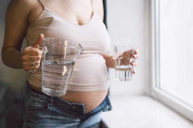 Acqua potabile della donna incinta vicino alla finestra. Maternità, concetto di gravidanza