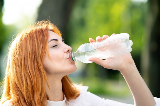 Acqua potabile della giovane donna assetata della testarossa da una bottiglia nel parco di estate.