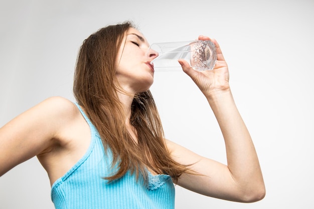 Acqua potabile della giovane donna da un bicchiere contro un muro bianco