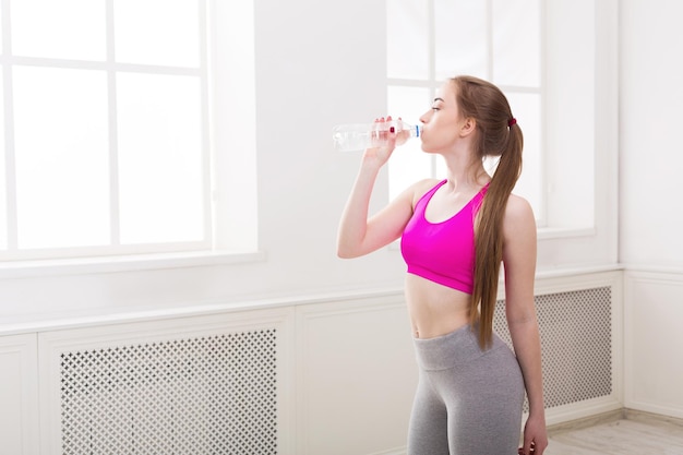 Acqua potabile della giovane donna di sport. Ragazza sicura di forma fisica che tiene bottiglia di plastica, riposo dopo l'allenamento, concetto di stile di vita sano