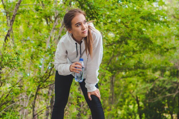 Acqua potabile della giovane ragazza caucasica felice stanca in parco mentre pareggiando. Copia spazio