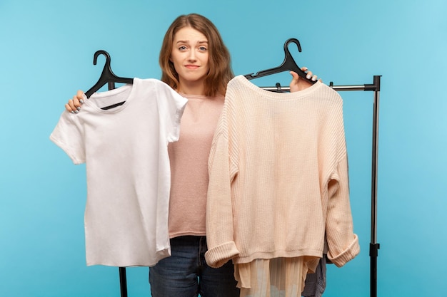 Acquirente donna confusa e insicura che tiene i vestiti sugli scaffali e guarda con dubbi pensando e scegliendo abiti alla moda che acquistano nella boutique moderna non può prendere una decisione girato in studio al coperto isolato