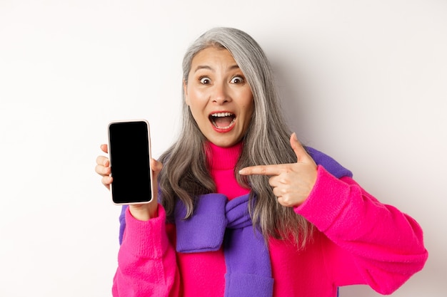 Acquisti online. Bella nonna asiatica sorridente, puntando il dito sullo schermo vuoto dello smartphone, guardando stupita, mostrando l'applicazione mobile, in piedi su sfondo bianco.