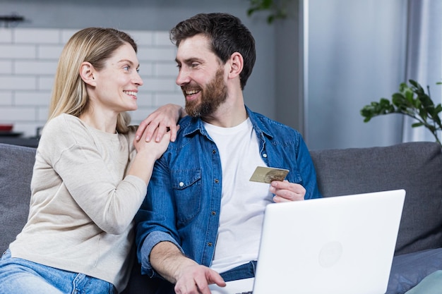 Acquisti online Famiglia uomo e donna a casa seduti sul divano che acquistano beni online da un laptop con in mano una carta di credito Abbracci sorridenti felici