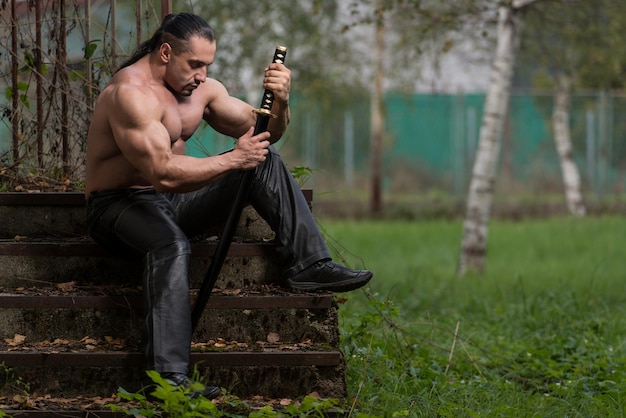 Action Hero muscoloso uomo che tiene una spada antica in piedi nella foresta che indossa pantaloni di pelle