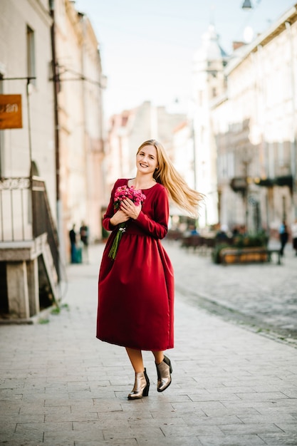 Adatti il ritratto di giovane donna alla moda della signora con il mazzo di fiori che cammina per strada, che porta vestito alla moda carino, bella ragazza in un vestito rosso che sorride gode dei suoi fine settimana.