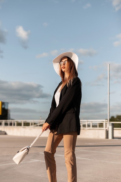 Adatti la giovane donna in vestiti alla moda casuali eleganti con la borsa dell'annata e il cappello di paglia posa sull'asfalto contro il cielo soleggiato blu. Elegante modello di ragazza elegante piuttosto voga in abiti giovanili in città.