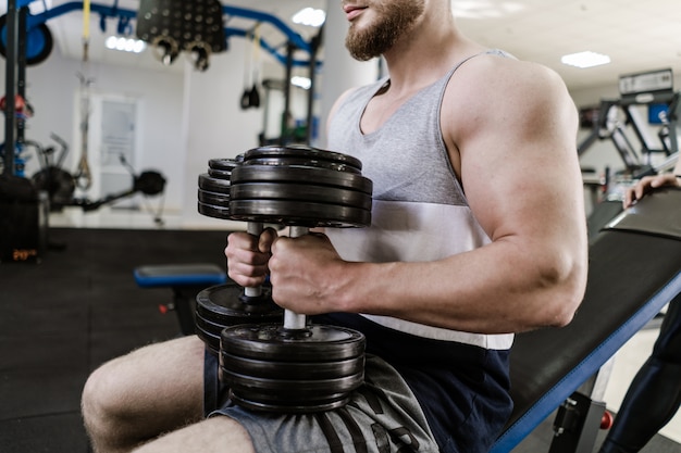 Addestramento muscolare bello dell'uomo con la testa di legno pesante in palestra. Il giovane con il grande bicipite si siede e fa allenamento in casa. Concetto di sport e salute.