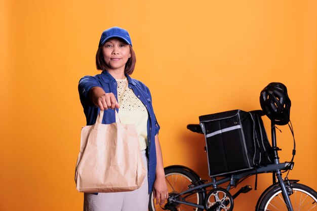 Addetto alle consegne in pizzeria che indossa l'uniforme blu mentre consegna l'ordine di fast food al cliente durante l'ora di pranzo. Impiegato del ristorante in piedi in studio con sfondo giallo. Concetto di servizio da asporto