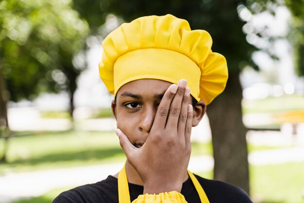 Adolescente africano nero cuoco con cappello da chef e grembiule giallo uniforme nascondere il viso con la mano Pubblicità creativa per bar o ristorante