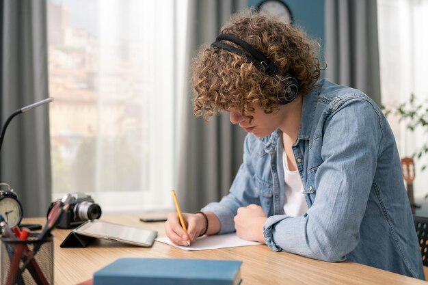 Adolescente americano concentrato che indossa le cuffie che scrivono le note studiano con il computer portatile ed i libri