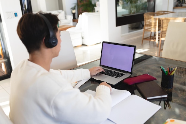 Adolescente asiatico che indossa le cuffie studiando su un computer portatile con libri sul tavolo a casa, copia spazio