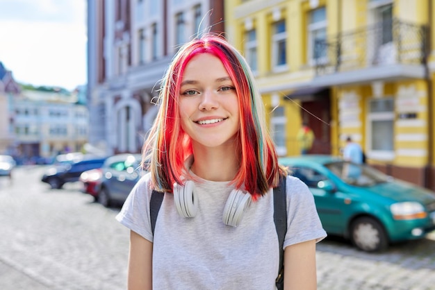 Adolescente bella ragazza alla moda 16, 17 anni in cuffie wireless con acconciatura colorata brillante sulla strada della città soleggiata estiva che guarda l'obbiettivo. Stile di vita, gioventù, moda, bellezza