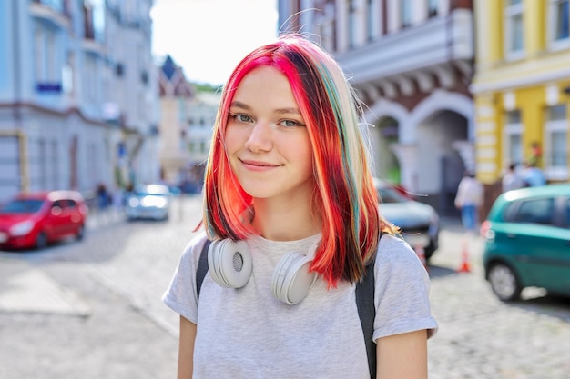 Adolescente bella ragazza alla moda 16, 17 anni in cuffie wireless con acconciatura colorata brillante sulla strada della città soleggiata estiva che guarda l'obbiettivo. Stile di vita, gioventù, moda, bellezza