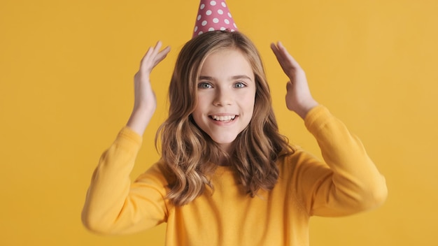 Adolescente bionda allegra in cappello del partito che posa felicemente sulla macchina fotografica che celebra il suo compleanno sopra priorità bassa gialla. Festeggiata