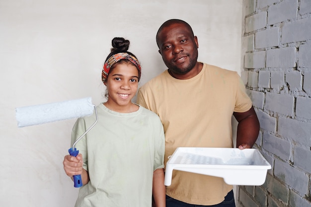 Adolescente carina felice con rullo di vernice e suo padre sorridente con contenitore di plastica quadrato bianco con vernice in piedi nell'angolo