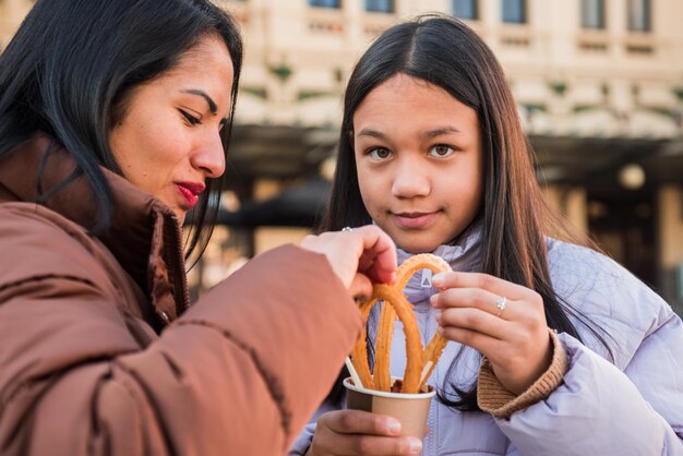 Adolescente che mangia spuntino guardando la macchina fotografica con sua madre Immergendo churros in una tazza di cioccolato