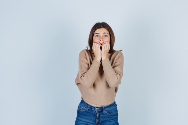 Adolescente che morde le unghie emotivamente in maglione, jeans e sembra spaventata. vista frontale.