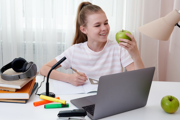 Adolescente che tiene mela verde che studia a casa con il computer portatile