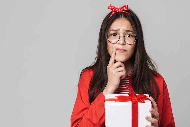 Adolescente chiedendosi carino che indossa un abito casual in piedi isolato su un muro grigio, con in mano una scatola regalo