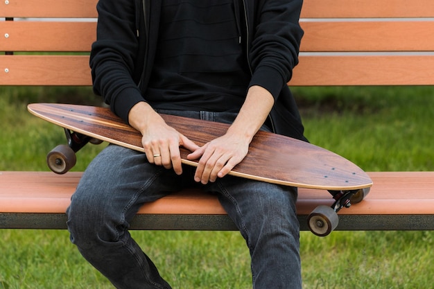 Adolescente dell'uomo che riposa e che si siede con lo skateboard o il longboard sulla panca di legno