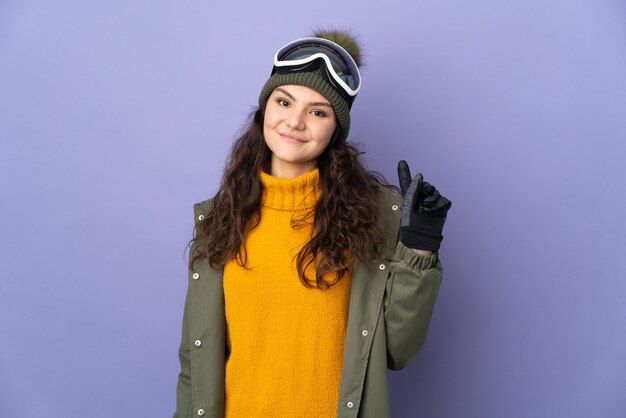 Adolescente donna russa con occhiali da snowboard isolati sulla parete viola che mostra e alzando un dito in segno del meglio