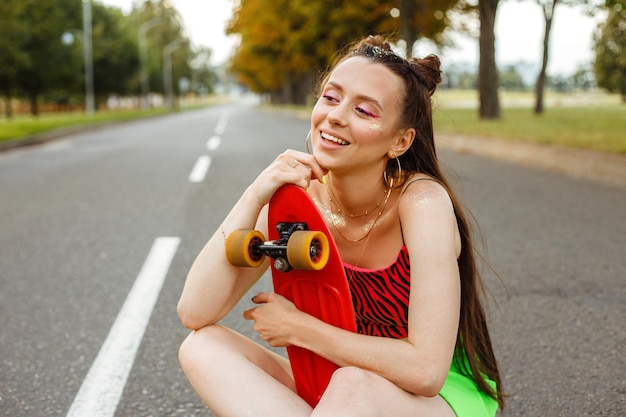 Adolescente felice con lo skateboard e in abito estivo alla moda seduto su strada in città