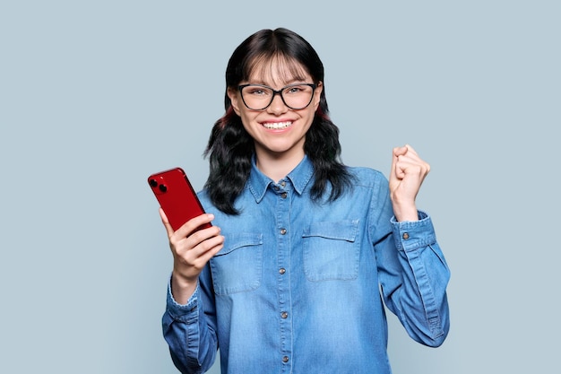 Adolescente felice con lo smartphone che fa il gesto del vincitore Adolescente femminile emozionale che tiene il pugno in su simbolo sì su sfondo di studio di colore grigio chiaro Gioia felicità emozione espressione stile di vita