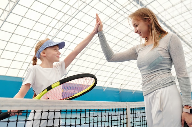 Adolescente felice in activewear che tiene la racchetta da tennis mentre dà il cinque al suo allenatore sopra la rete dopo aver giocato con successo sullo stadio moderno