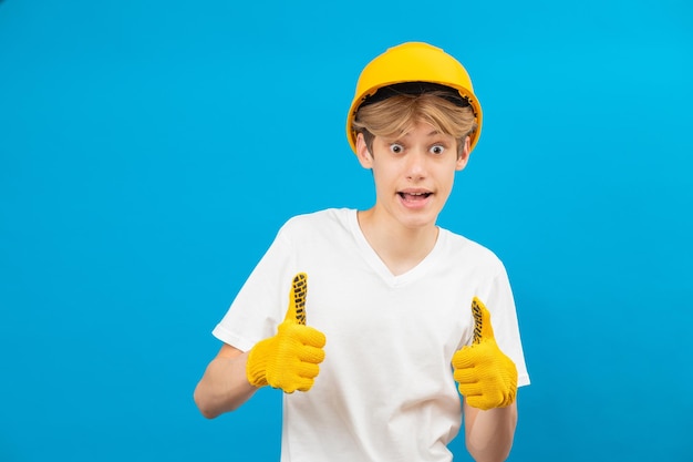 Adolescente felice in maglietta bianca e guanti in mano con casco giallo in piedi in studio su sfondo blu Un futuro architetto isolato su sfondo blu