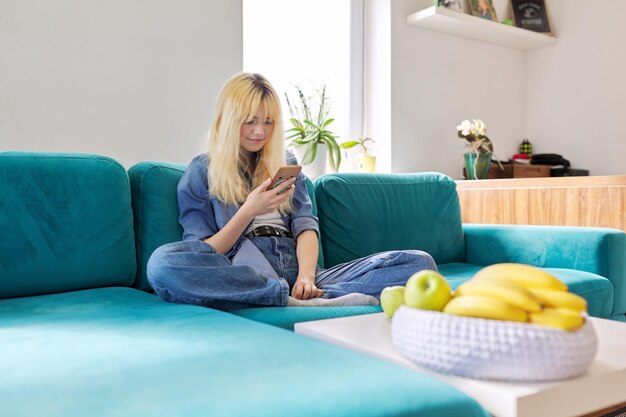 Adolescente felice sorridente seduto a casa sul divano guardando lo schermo dello smartphone