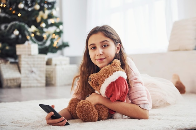 Adolescente femmina carina sdraiata al chiuso nel soggiorno durante le vacanze con telefono e orsacchiotto.