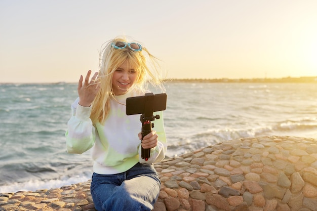 Adolescente femminile con smartphone su treppiede sulla spiaggia al tramonto sul mare che registra video